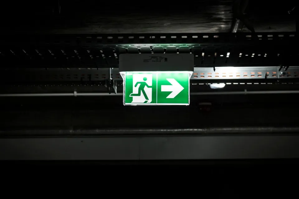 Illuminated green running-person exit sign with a right arrow beneath cable trays
